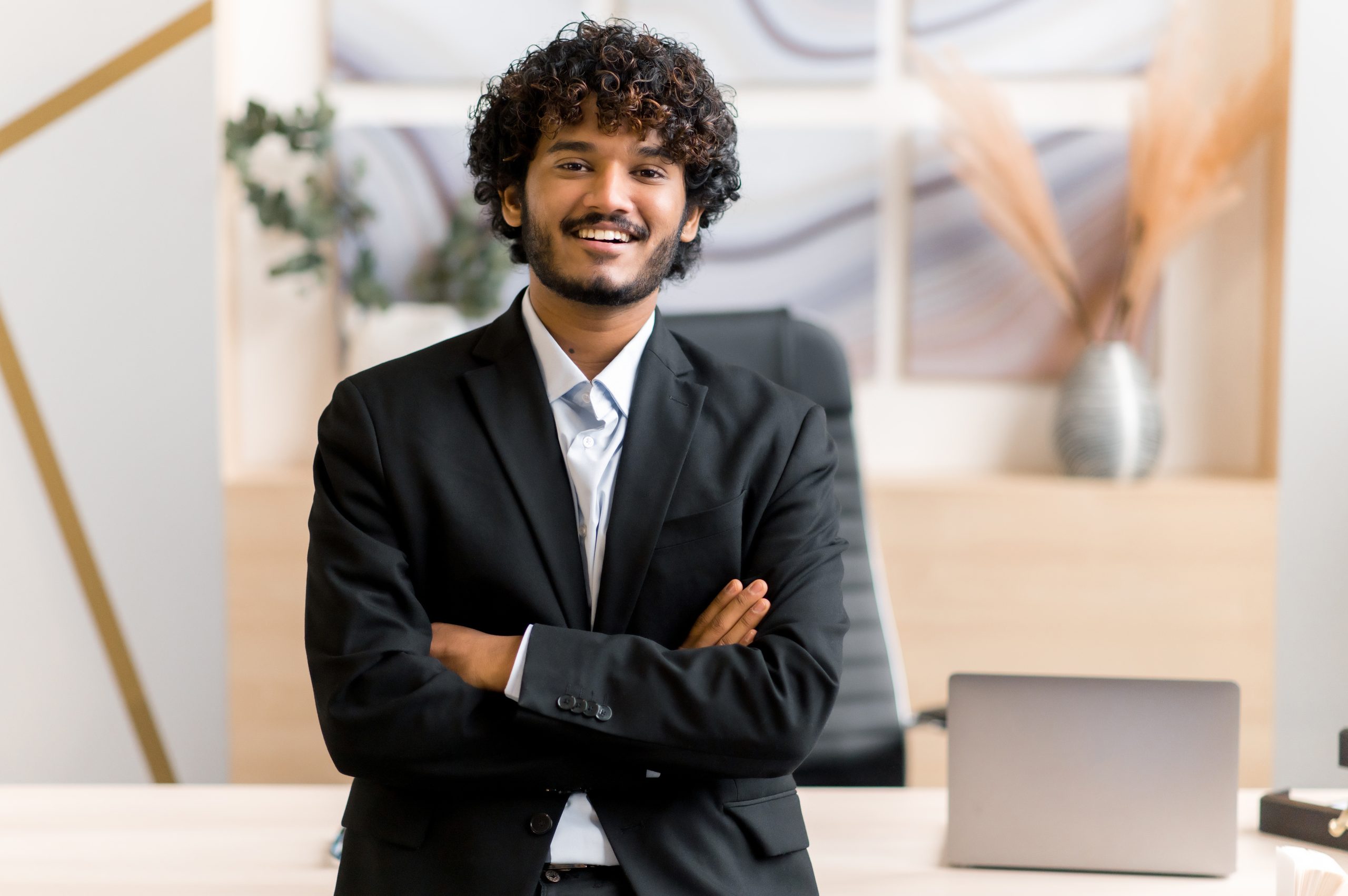 Portrait successful attractive young adult Indian or Arabian business leader, manager or IT professional dressed in formal suit stand near desk in office with arms crossed, look at camera, smile
